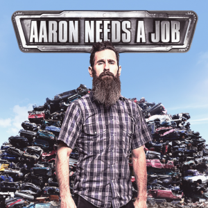 Aaron Needs a Job - A man with a long beard stands in front of a junkpile of cars,