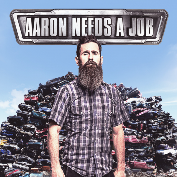 Aaron Needs a Job - A man with a long beard stands in front of a junkpile of cars,